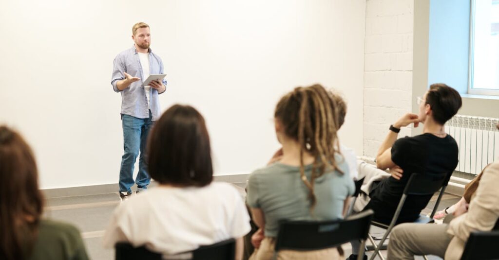 Man presenting with tablet to a small seated audience in a modern indoor setting.